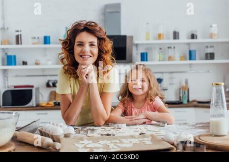 jeune mère et fille regardant l'appareil photo près de biscuits crus et les ingrédients sur la table de cuisine Banque D'Images