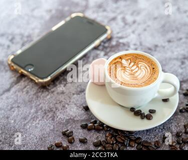 Café sur un mug blanc avec une soucoupe avec Marshmallow, grains de café et flou de fond de téléphone mobile Banque D'Images