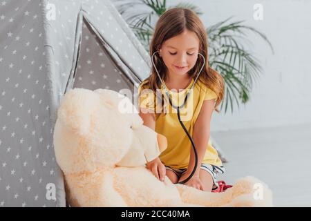 fille en t-shirt jaune jouant docteur avec stéthoscope et teddy porter une tente de jeu proche Banque D'Images