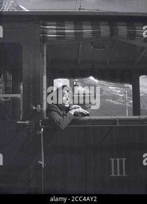 1950s, historique, une femme assise à l'intérieur d'une calèche suisse, penchée d'une fenêtre ouverte, Alpes suisses, Suisse. Le numéro 3 en chiffres romains est en lettres sur la porte de la gare. Banque D'Images