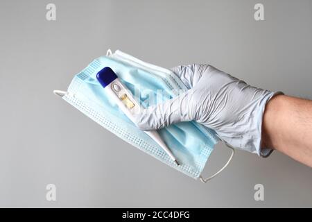 les mains du médecin protégées par des gants bleus tiennent un masque chirurgical bleu et un thermomètre anti-fièvre sur fond gris, contre la pandémie. Banque D'Images