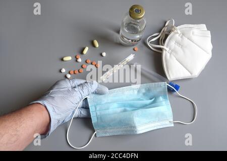 la main du médecin protégée par des gants bleus tient un masque chirurgical et un thermomètre anti-fièvre sur fond gris avec des masques ffp2 et des pilules de médicaments. Banque D'Images