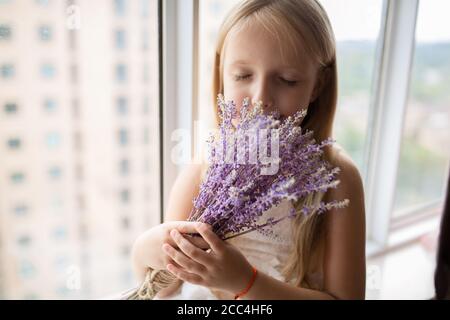 Jolie petite fille caucasienne avec cheveux blonds tenant un bouquet de fleurs violettes à la maison. Enfant debout près de la fenêtre pendant la pandémie de covid-19 du coronavirus Banque D'Images