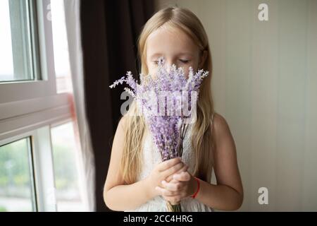 Jolie petite fille caucasienne avec cheveux blonds tenant un bouquet de fleurs violettes à la maison. Enfant debout près de la fenêtre pendant la pandémie de covid-19 du coronavirus Banque D'Images