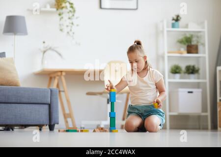 Fille avec le syndrome de Down assise sur le sol dans le chambre et construction d'une tour à partir de blocs de couleurs Banque D'Images
