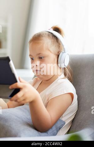 Petite fille avec le syndrome de Down portant des écouteurs et regardant des dessins animés sur une tablette numérique tout en étant assis sur un canapé dans la chambre Banque D'Images