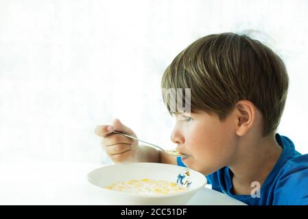 Un garçon dans un T-shirt bleu a le petit déjeuner avec des flocons d'avoine et du lait dans une assiette blanche, espace pour le texte, isoler Banque D'Images