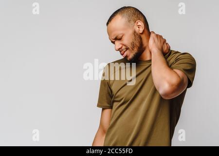 jeune afro-américain en t-shirt vert sur fond gris sensation d'inconfort physique pour les douleurs au cou massant les muscles tendus Banque D'Images