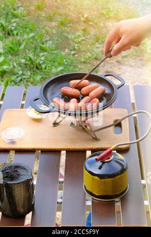 Camping fabrication de nourriture. Saucisses sur la poêle sur le brûleur à gaz touristique. Campeur préparant la nourriture dans la forêt. Banque D'Images