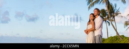 Couple relaxant regardant le coucher du soleil sur la bannière de vacances à la plage. Les gens multiraciaux appréciant les vacances tropicales ensemble enserrant dans l'amour regarder loin Banque D'Images