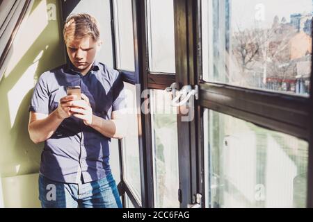 Jeune homme avec téléphone portable rester près de la fenêtre dedans lumière de jour contrastée Banque D'Images