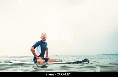 L'homme surfeur est assis sur une planche de surf dans l'eau et attend les vagues Banque D'Images