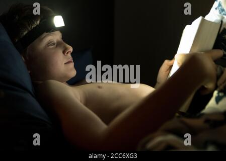 Lecture au lit à la nuit concept photo. Petit garçon de lecteur utilise la torche de tête pour un livre lisant dans le lit Banque D'Images