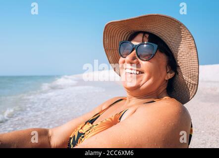 Bonne femme âgée souriante dans des lunettes de soleil et un grand chapeau prendre un bain de soleil en bord de mer Banque D'Images