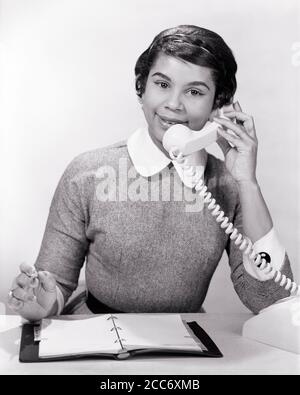 ANNÉES 1950, UNE FEMME AFRO-AMÉRICAINE JOYEUSE AU BUREAU PARLANT AU TÉLÉPHONE CARNET DE RENDEZ-VOUS À ADRESSE OUVERTE SOURIANT REGARDANT LA CAMÉRA - N115 HAR001 HARS 1 RÉCEPTIONNISTE COMMUNICATION FACIALE CARRIÈRE JEUNES ADULTES INFORMATION ASSISTANT SATISFAIT JOIE STYLE DE VIE SATISFACTION COMMIS FEMMES EMPLOIS STUDIO SHOT SANTÉ MAISON VIE COMMUNIQUER ESPACE DE COPIE DEMI-LONGUEUR FEMMES PERSONNES PROFESSION EXPRESSIONS DE CONFIANCE B&W CONTACT VISUEL COMPÉTENCE PROFESSION BONHEUR COMPÉTENCES ADRESSE JOYEUX SERVICE À LA CLIENTÈLE AFRO-AMÉRICAINS RÉSEAU AFRO-AMÉRICAIN CARRIÈRES MAIN-D'ŒUVRE ETHNIQUE NOIRE OPPORTUNITÉ DE NOMINATION D'UN EMPLOYÉ DU BUREAU DE FIERTÉ Banque D'Images