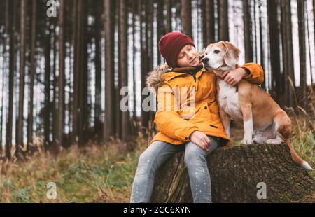 Portrait de deux meilleurs amis qui s'embrasent - garçon et son chien de beagle s'assoient sur la souche de l'arbre dans la forêt de pins d'automne. Image du concept humain et animal domestique. Banque D'Images