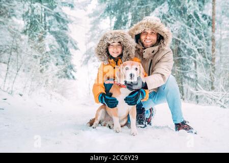 Père et fils vêtus de chaud capuche décontracté Parka Veste Outerwear marchant avec leur chien de beagle dans la forêt enneigée gai souriant visages portrait. PE Banque D'Images