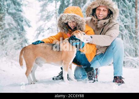 Père et fils; vêtus de chaud capuche décontracté Parka Veste Outerwear marchant avec leur chien de beagle; ils le petent et souriant dans la forêt enneigée . Animaux de compagnie Banque D'Images