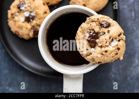 Un biscuit tahini avec des morceaux de chocolat mis sur le bord d'une tasse en céramique blanche remplie de café foncé sur une assiette noire. Banque D'Images