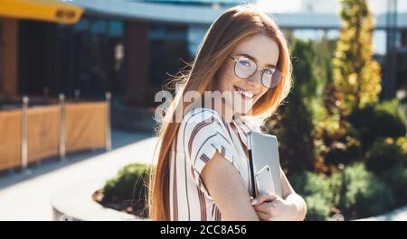 Freckled femme caucasienne avec des cheveux rouges et des lunettes embrassant un ordinateur portable et posé à la caméra avec un téléphone Banque D'Images