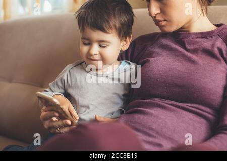 Femme enceinte de race blanche et son fils utilisant un mobile blanc asseyez-vous sur le canapé et souriez Banque D'Images