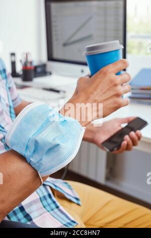 gros plan sur un jeune homme du caucase assis à son bureau bureau portant son masque facial dans son bras pendant qu'il est boire un café Banque D'Images