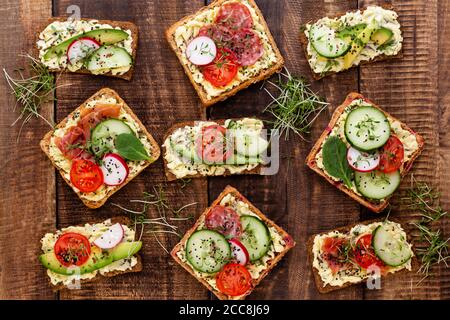 Variété de mini sandwiches avec fromage à la crème, légumes et salami. Sandwichs au concombre, radis, tomates, salami sur fond gris, vue du dessus Banque D'Images