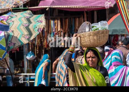 Orcha, Madhya Pradesh, Inde - Mars 2019: Une femme indienne portant un panier en bois avec des ustensiles en acier sur sa tête dans le marché coloré dans le ci Banque D'Images