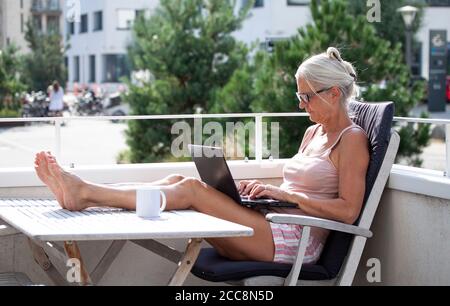 Femme sénior pieds nus assise et dactylographiant sur un ordinateur portable ordinateur à l'extérieur sur un balcon dans un complexe résidentiel moderne les pieds vers le haut Banque D'Images