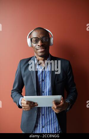 Portrait d'un jeune homme africain en lunettes et casque sans fil sourire à l'appareil photo tout en utilisant le tablet pc contre le marron arrière-plan Banque D'Images