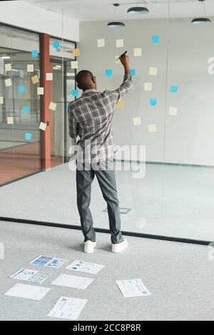 Vue arrière de l'homme d'affaires africain debout devant le verre tableau noir et notes sur les notes adhésives au bureau Banque D'Images