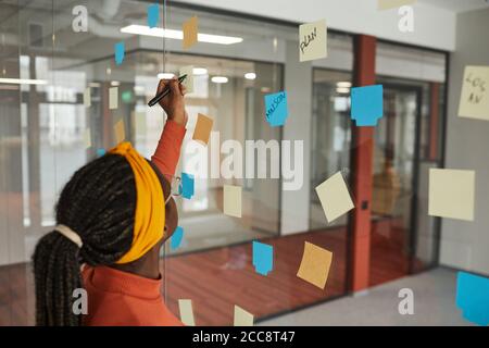 Vue arrière de la femme africaine avec belle coiffure l'écrivant idées sur les notes adhésives sur le mur de verre au bureau Banque D'Images