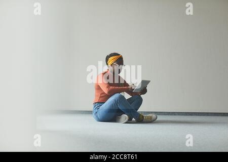 Jeune femme africaine assise sur le sol dans une pièce vide et à l'aide d'une tablette numérique, elle travaille en ligne Banque D'Images