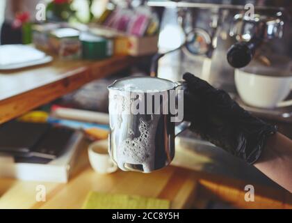 Barista portant des gants médicaux noirs en latex, préparant une boisson à l'aide d'une machine à espresso dans le café du magasin. Une femme tient le pichet de lait en métal à la main. Banque D'Images