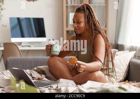Jeune femme avec belle coiffure assis sur le lit regarder un film sur ordinateur portable et prendre le petit déjeuner dans la chambre Banque D'Images
