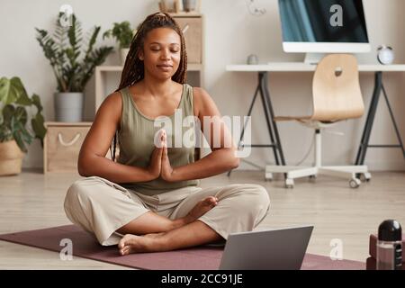 Jeune femme assise en position lotus sur un tapis d'exercice avec ses yeux fermés et méditant dans le salon Banque D'Images