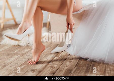 Vue courte de la mariée mettant sur les talons de mariage près de la robe sur le lit Banque D'Images