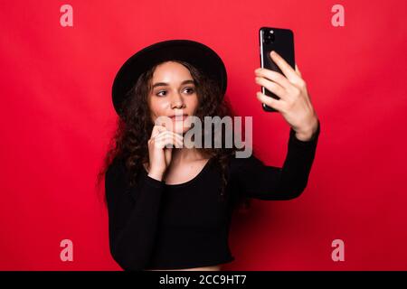 Gros plan portrait belle femme prendre selfie sur l'appareil photo avant de smartphone moderne isolé sur fond rouge brillant Banque D'Images