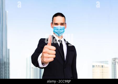 Bravo ! Jeune homme d'affaires donnant l'approbation de mouvement de main vers l'appareil photo portant un masque médical dans la ville. Banque D'Images