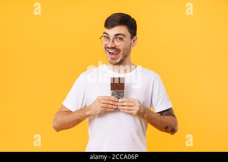 Photo du jeune homme heureux dans les lunettes tenant le chocolat et lécher ses lèvres isolées sur fond jaune Banque D'Images