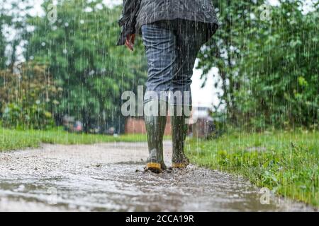 Vue arrière à angle bas de la femme (partie inférieure) portant des bottes wellington, des bottes en caoutchouc, sous une forte pluie en marchant loin de la caméra le long d'un sentier britannique boueux. Banque D'Images