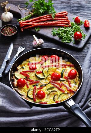 frittata, omelette avec courgettes, fines saucisses fumées et tomates remplissant dans une poêle sur une table en bois sombre avec des ingrédients à l'arrière-plan, vert Banque D'Images