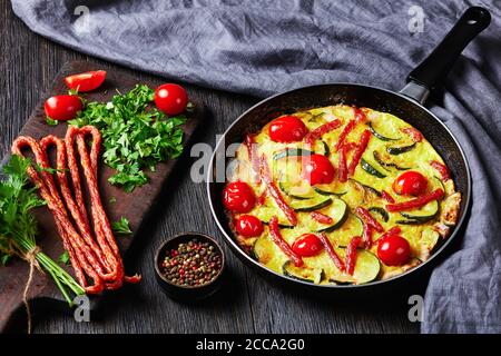 frittata, omelette avec courgettes, fines saucisses fumées et tomates remplissant dans une poêle sur une table en bois sombre, vue horizontale d'en haut, gros plan Banque D'Images