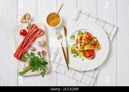 frittata, omelette avec courgettes, fines saucisses fumées et tomates remplissant sur une plaque blanche sur une table en bois avec des ingrédients sur une planche à découper, horizontales Banque D'Images