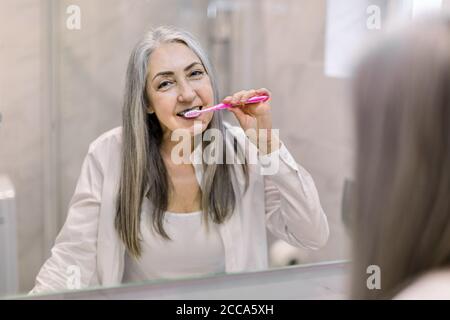 Reflet dans le miroir de la belle femme âgée souriante avec de longs cheveux gris droits, debout dans la salle de bains, se brossant les dents et appréciant son matin Banque D'Images