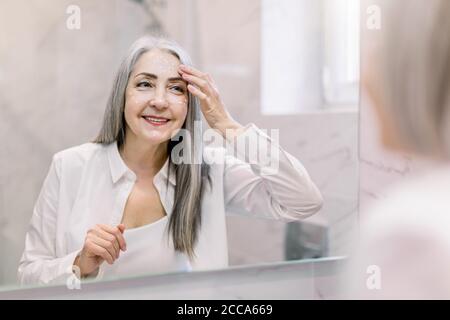 Jolie femme âgée avec de longs cheveux gris, portant une chemise blanche, regardant son visage dans le miroir de salle de bains, et appliquant de la crème anti-rides ou cosmétique Banque D'Images