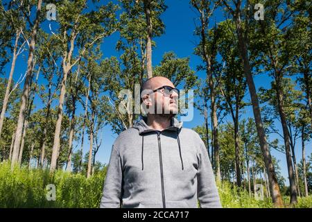 Homme adulte à capuche et lunettes de soleil marchant dans la forêt verte le beau jour d'été, portrait à moitié de corps, angle bas, concept de randonnée Banque D'Images