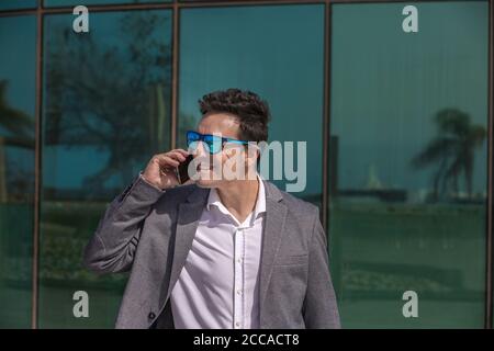 Joyeux homme d'affaires adulte réussi en tenue habillée et lunettes de soleil tendance avoir une conversation téléphonique tout en se tenant contre le mur en verre réfléchissant de mode Banque D'Images