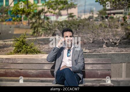 Homme d'affaires souriant, bien habillé, assis sur un banc en bois et discuter de nouvelles pendant un appel téléphonique tout en ayant une pause et repos sur la rue à l'intérieur Banque D'Images