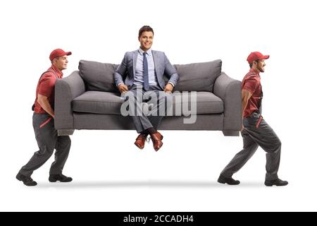 Homme souriant dans des vêtements élégants assis sur un canapé à maison et deux déménageurs transportant le canapé Banque D'Images